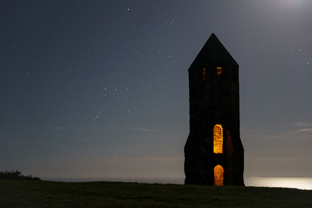 A nighttime photo from a hilltop overlooking the sea. A large stone tower is dominant on the right of the frame, shaped like a rocket with a near- conical roof. The presence of a door & window aperture within the tower has been revealed by the photographer adding warm orange light inside the structure which is otherwise rendered in silhouette. The constellation of Orion is visible in the night sky to the tower’s left and there’s a hint of moonlight reflecting on the distant water